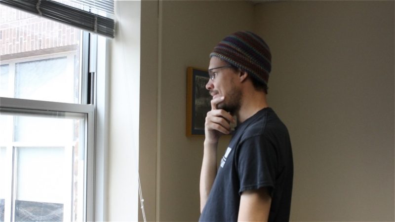 Greene wears a knitted cap and a black t-shirt and glasses while standing and looking out the window of a room in Shanks Hall.